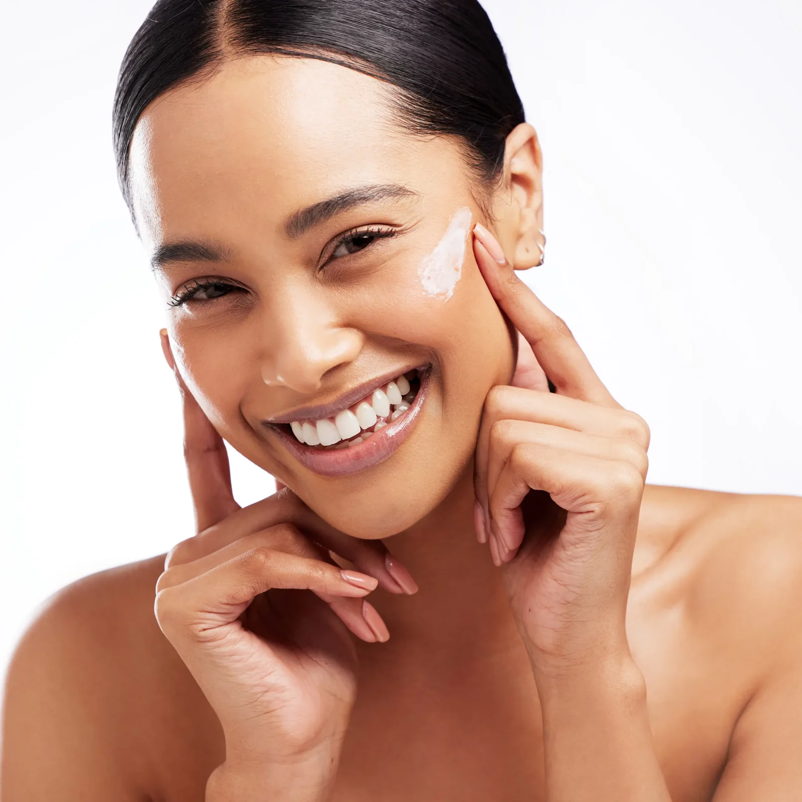 Close-up of a smiling woman applying white cream to her cheek with her fingers, demonstrating skincare routine.