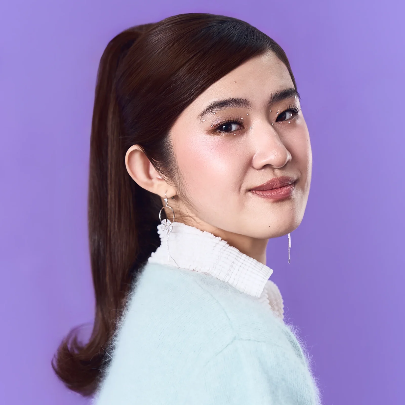 Close-up side profile of an Asian woman with rhinestone face decorations around the eye and cheek, wearing a white textured collar against a purple background.
