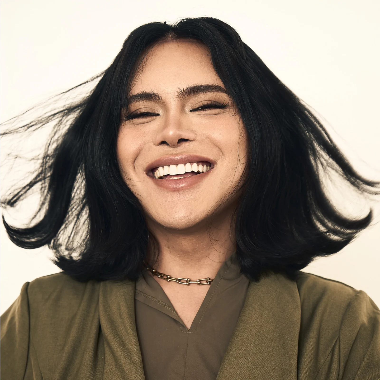 Close-up of a smiling woman with dark hair, olive jacket, and brown top against a light background.