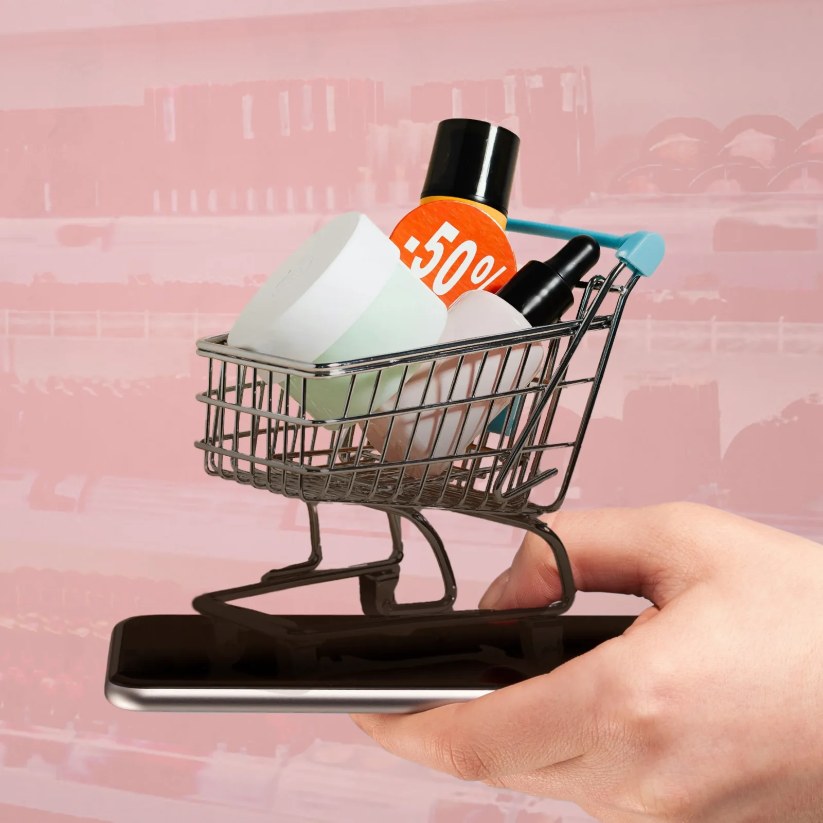 Hand holds a small shopping cart overflowing with products, including a bright 50% off tag, against a pink background.