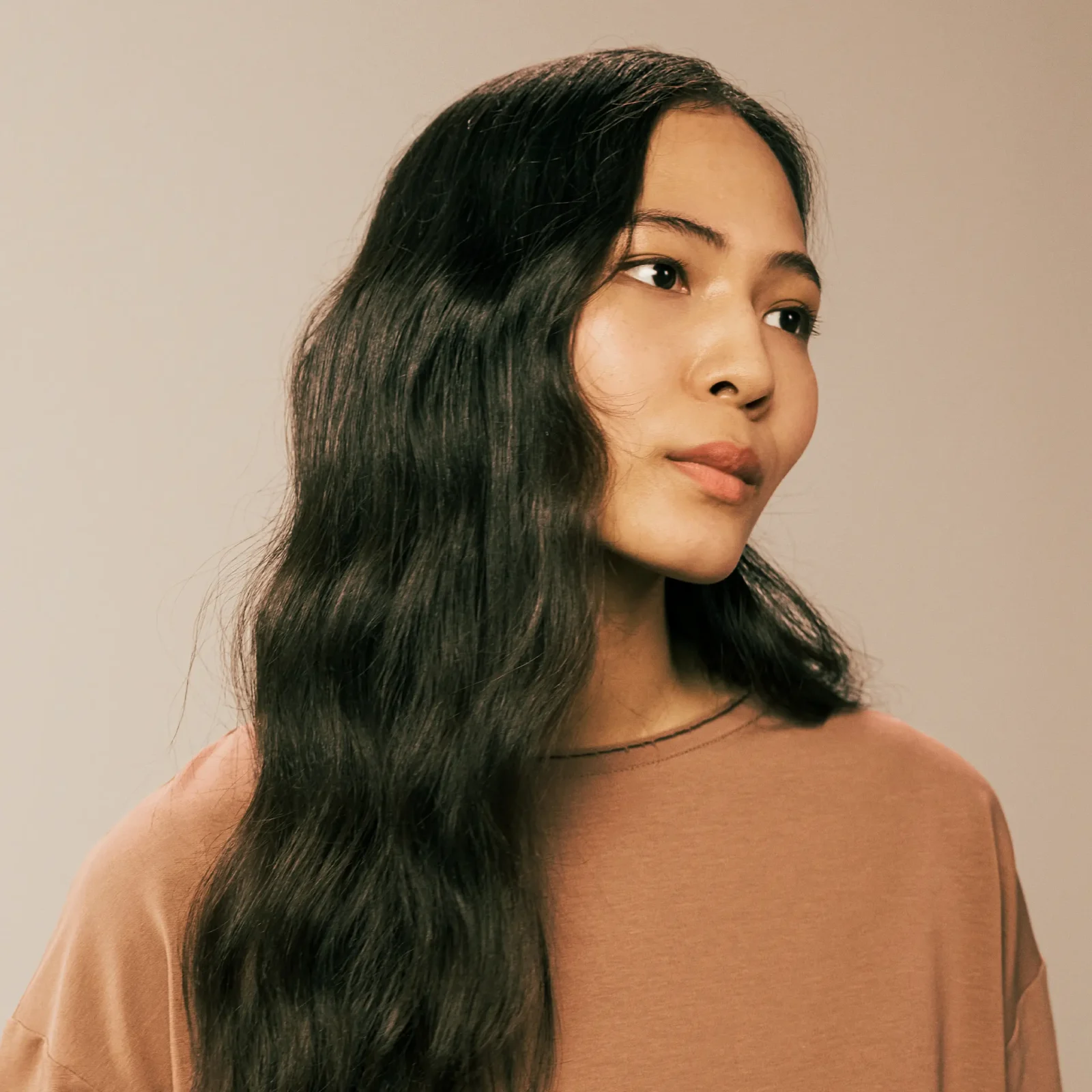 Close-up of a woman with long dark hair, wearing a peach top, against a beige background.
