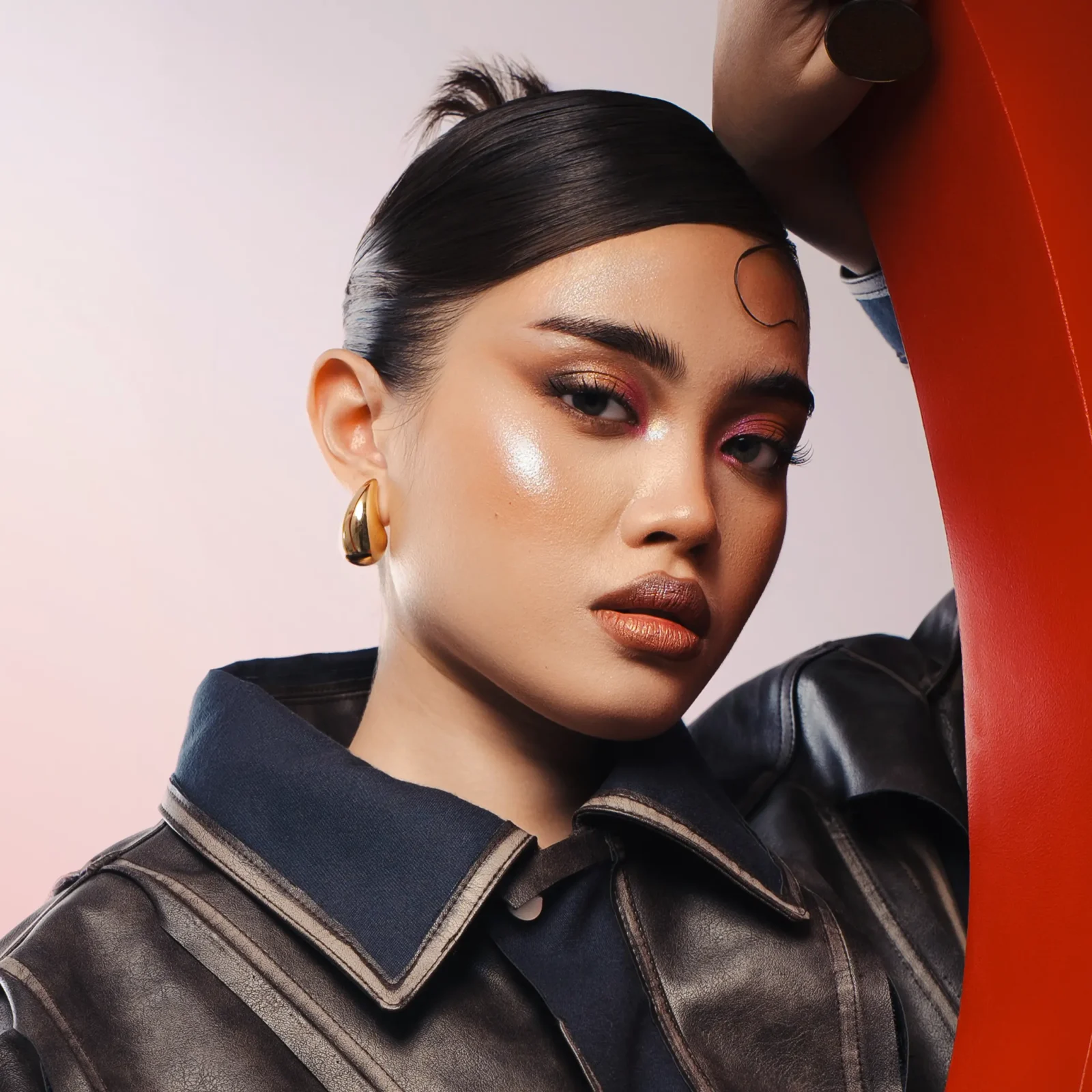 Close-up portrait of a woman with slicked-back hair, wearing a dark leather jacket and gold hoop earring, against a pink and orange background.