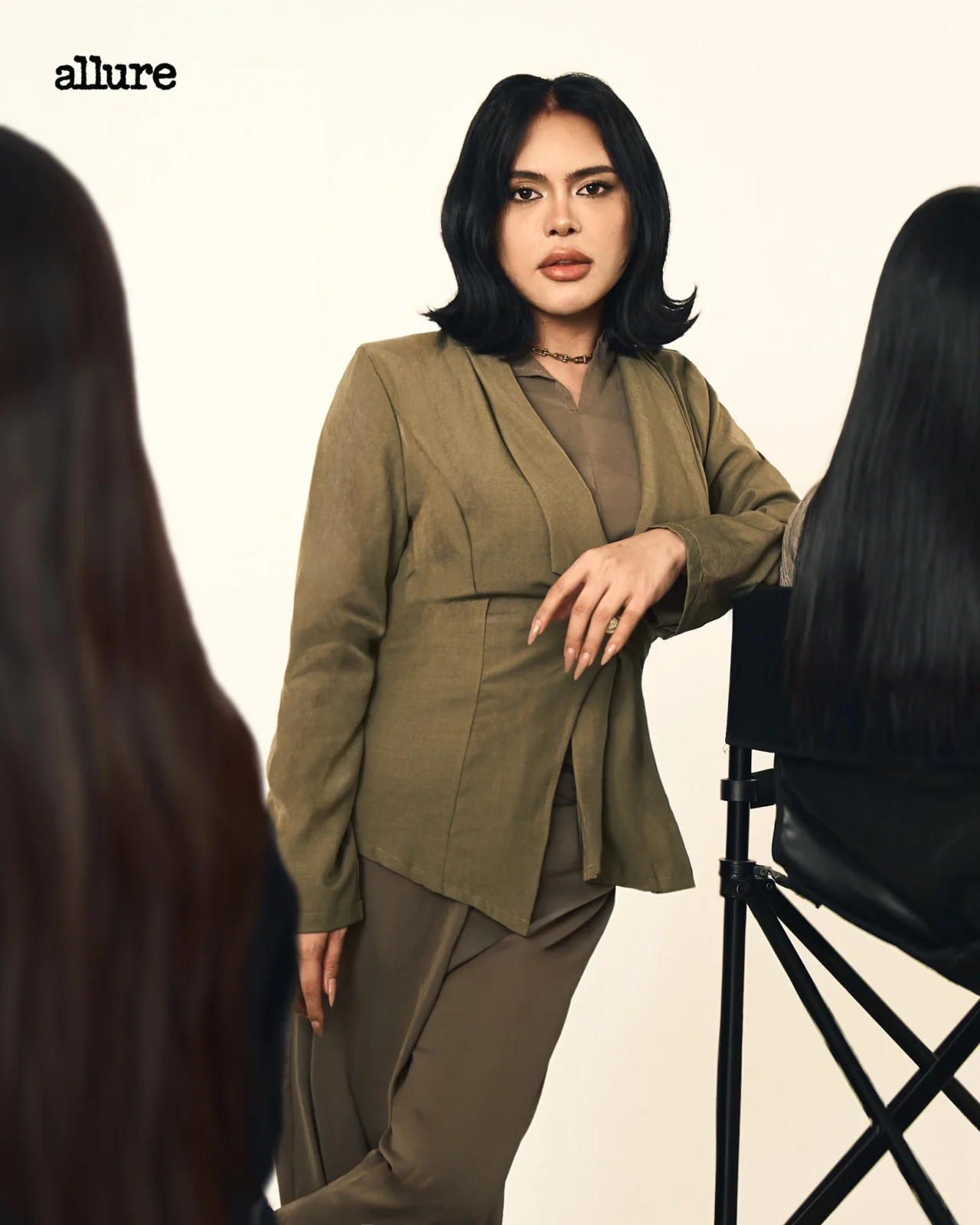 Woman in an olive blazer posing in a photography studio with the Allure logo visible in the upper left corner.