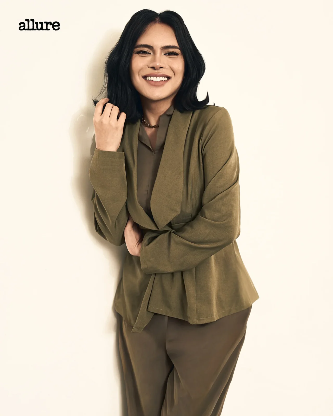 Portrait of a smiling woman with shoulder-length black hair, wearing an olive blazer, leaning against a light wall; Allure logo visible in the top-left corner.