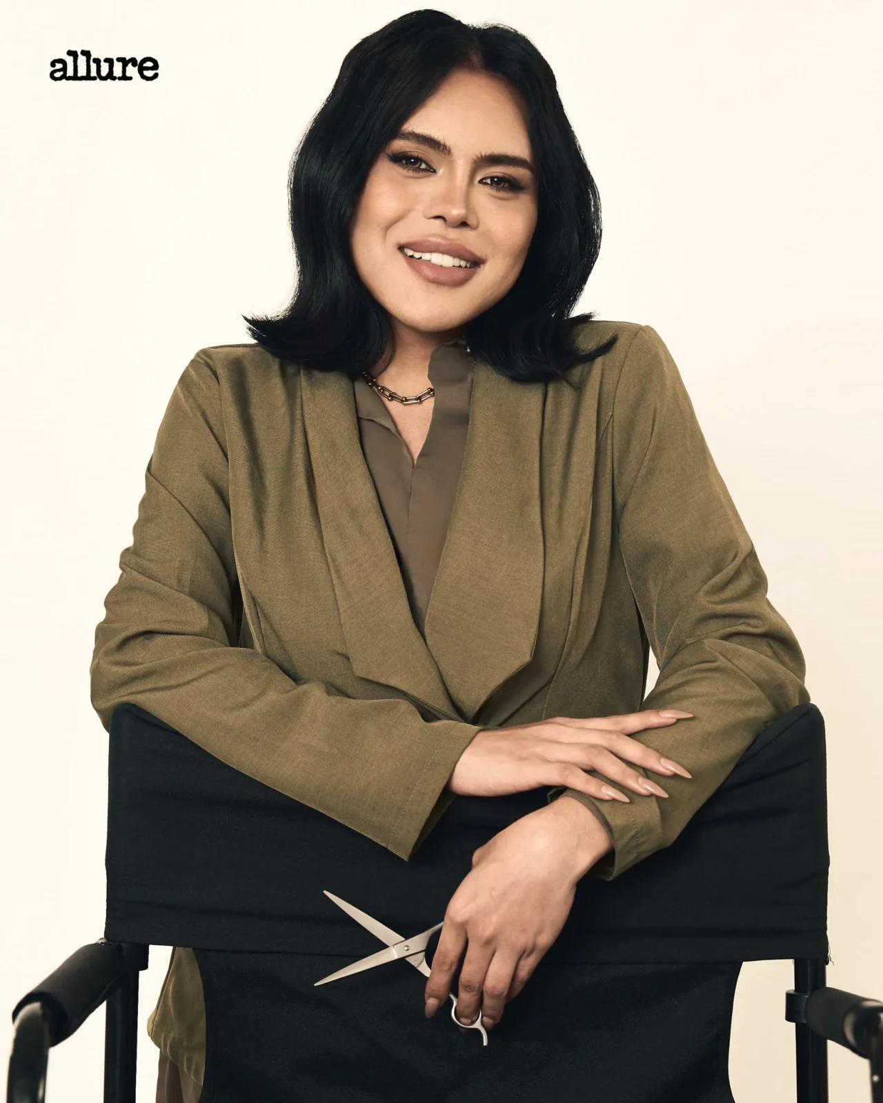 Woman wearing a tan blazer seated on a chair, hands resting on a large pair of scissors on her lap; Allure logo in the top-left corner.