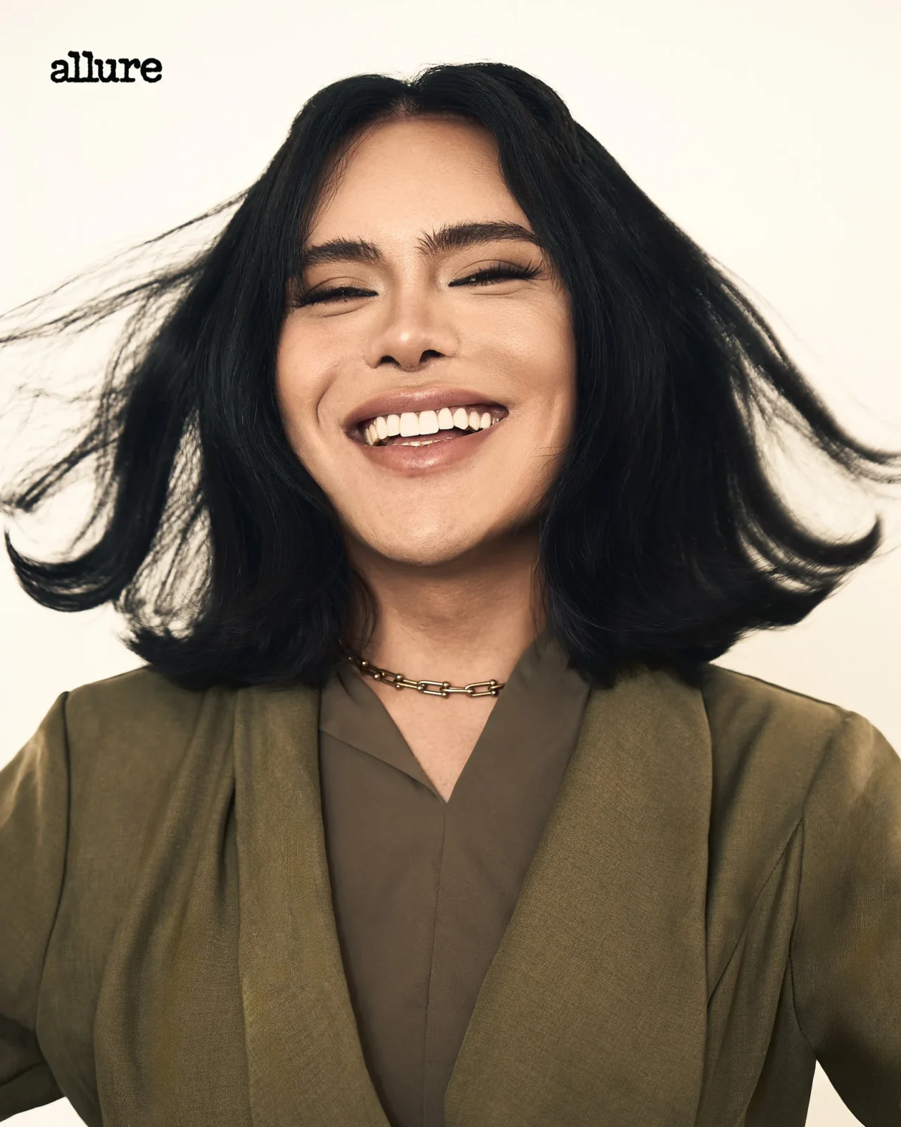 Close-up portrait of a smiling woman with shoulder-length black hair, wearing an olive blazer and a gold chain necklace. Allure text appears in the top-left corner.