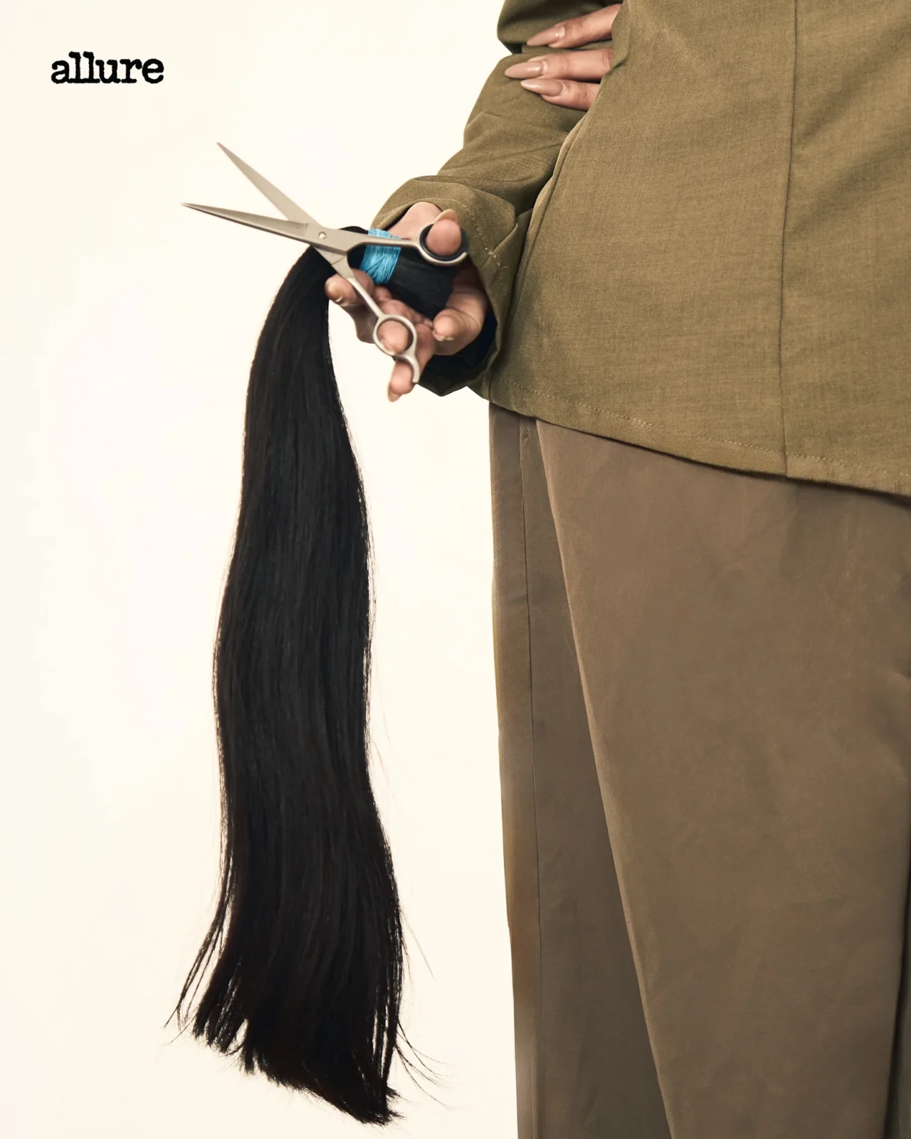 Person in a brown outfit cutting a long black ponytail with scissors, hair falling downward against a pale background; blue wrap around the wrist.