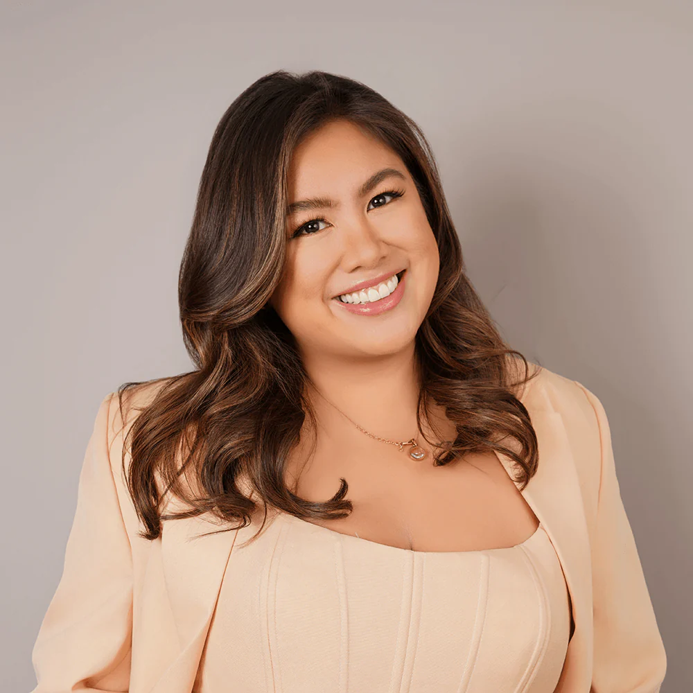 Smiling woman with wavy brown hair in a light peach outfit against a neutral gray background.