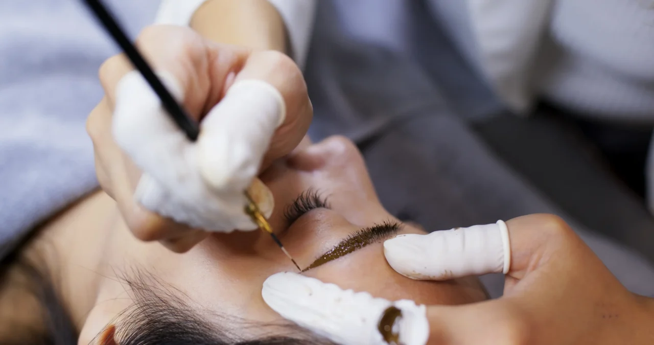Close-up of a cosmetic brow procedure: a technician in gloves deposits pigment along the client's eyebrow with a fine tool