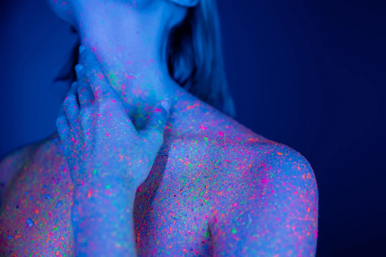 Close-up of a person's neck and shoulders covered in neon UV paint under blue blacklight (glow-in-the-dark speckles).