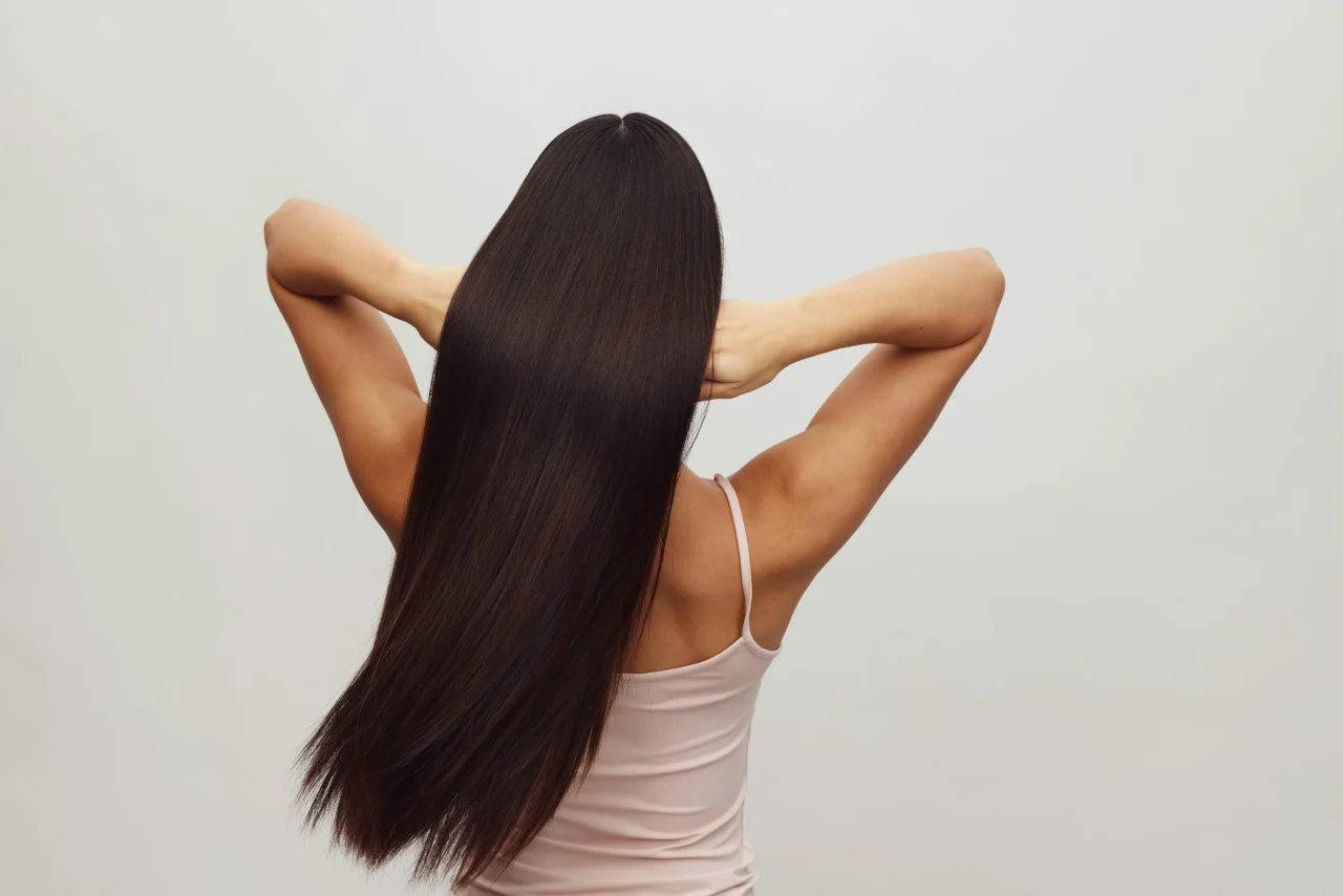 Person facing away with long straight dark hair, arms raised and hands behind the head, wearing a light pink tank top