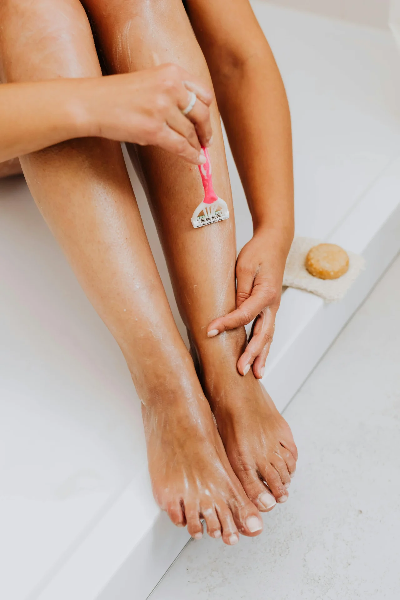 Person shaving their leg with a pink razor in a bathtub, applying shaved cream on smooth legs with a sponge nearby
