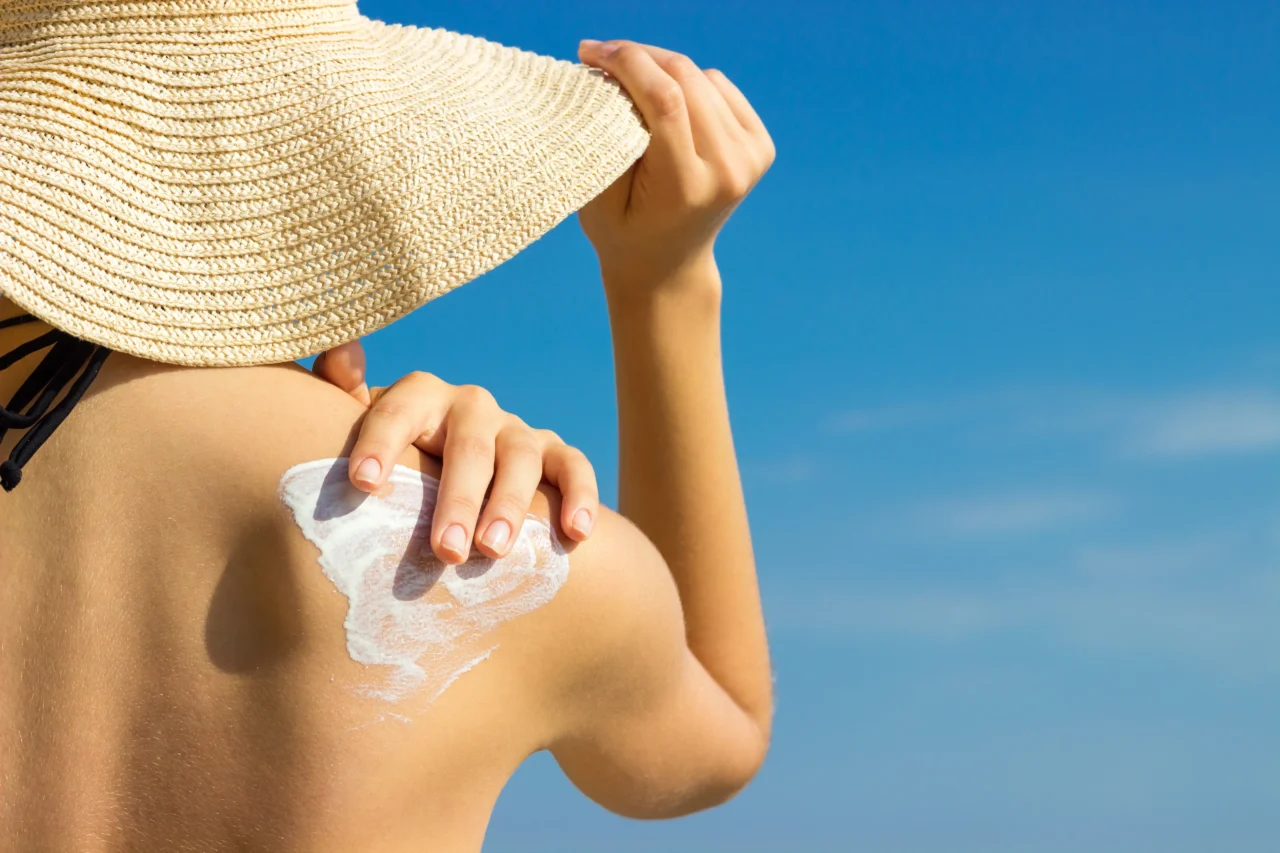Person applying sunscreen to their shoulder outdoors, with a straw hat and a clear blue sky behind them.