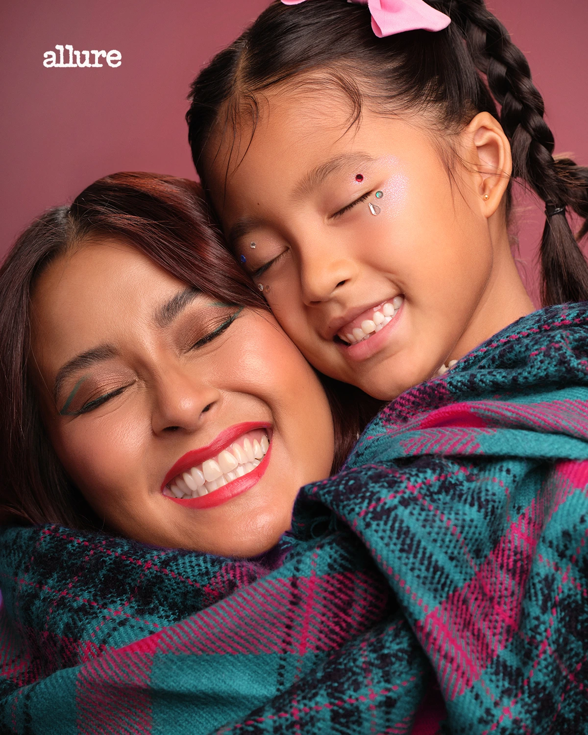 Woman and child hug, both smiling with eyes closed; a warm pink background and colorful blanket visible. Allure logo in corner appears.”