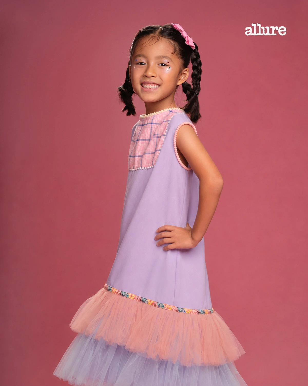 Young girl in a pastel lavender dress with pink tulle skirt, smiling against a pink studio backdrop; Allure logo in the corner.