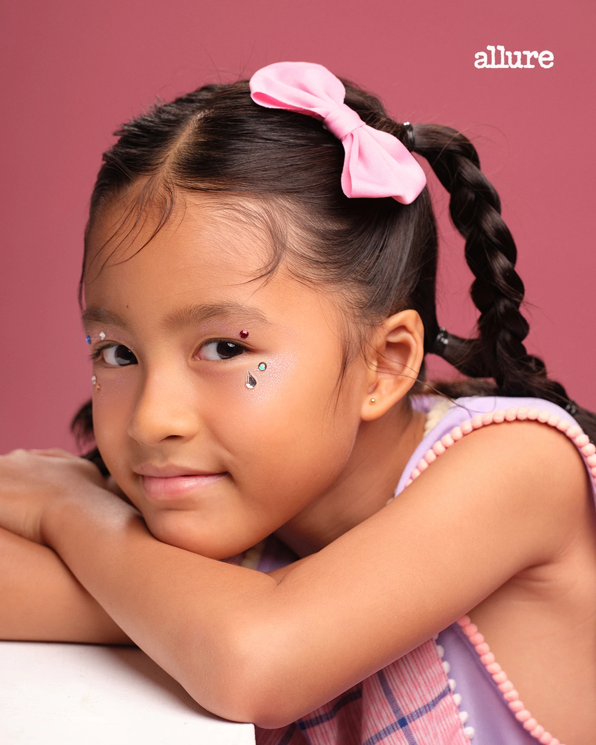 Young girl with braided hair and a pink bow rests her chin on her arms against a pink backdrop, wearing glitter stickers on her cheeks.