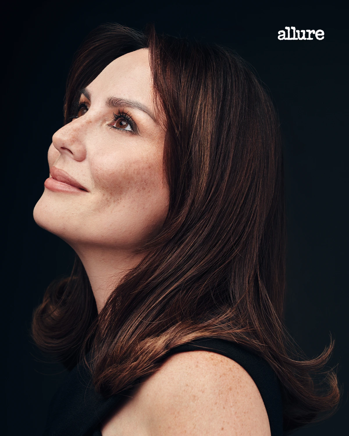 Portrait of a woman with brown hair looking upward to the left against a dark background, Allure logo in the corner.