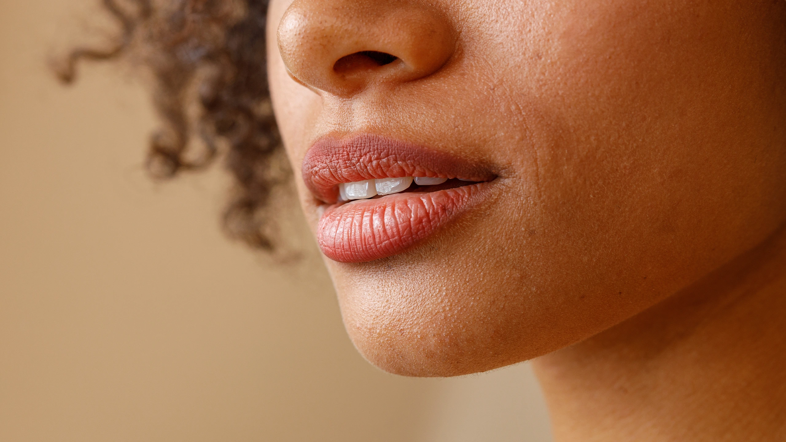 Close-up of a brown-skinned woman's lips and nose, lips slightly parted showing teeth, in soft lighting.