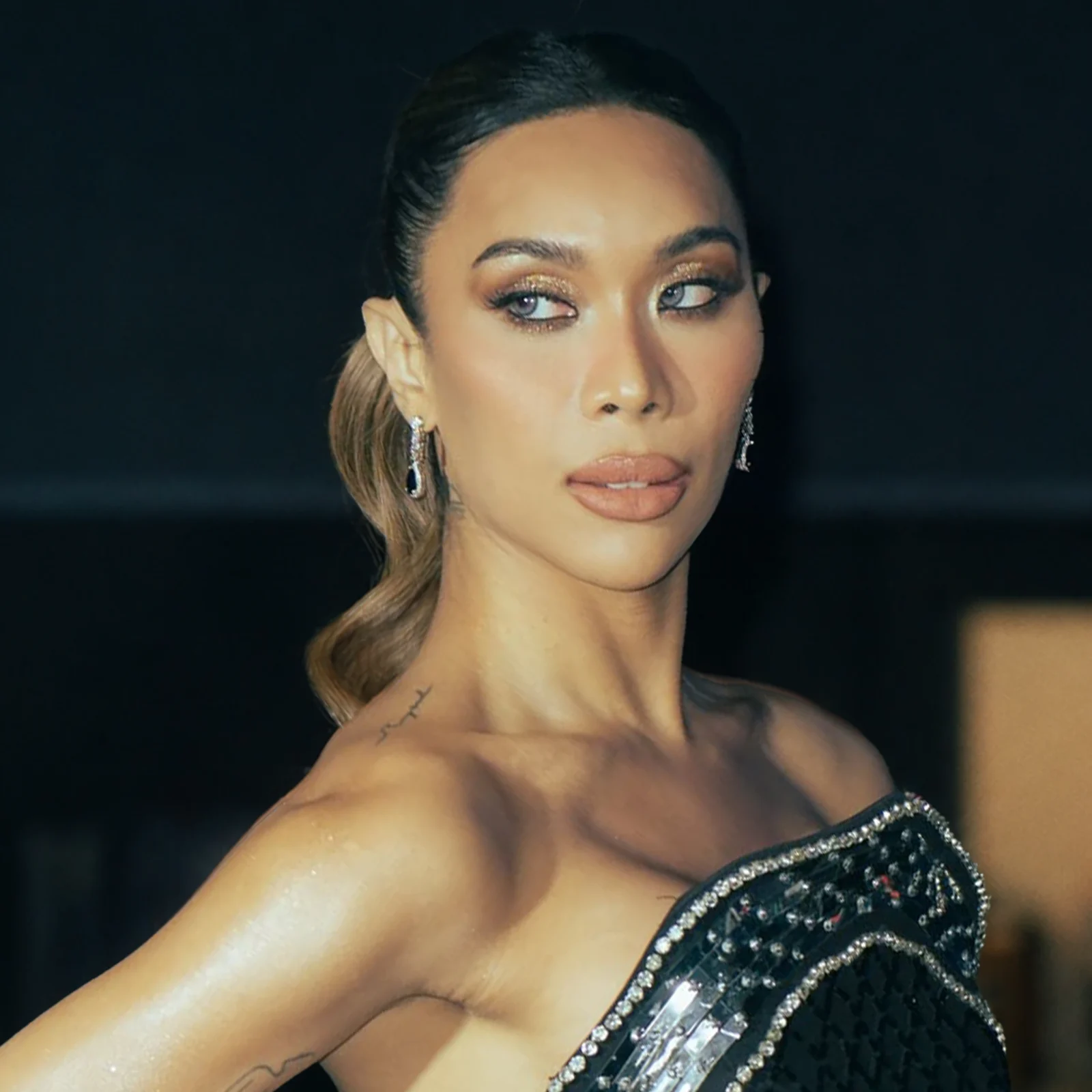 Close-up of a woman's face with glamorous makeup, diamond earrings, and a sequined black dress visible on the right.