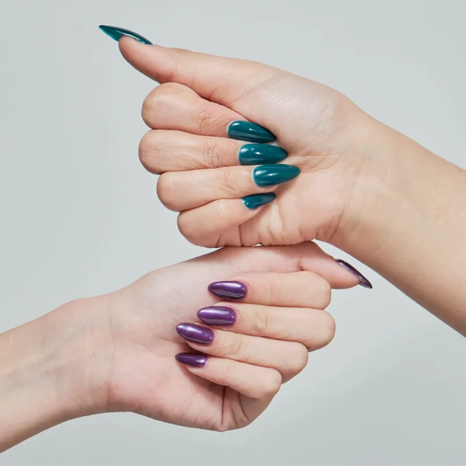 Close-up of hands with glossy teal and purple nail polish, interlaced against a pale gray background.