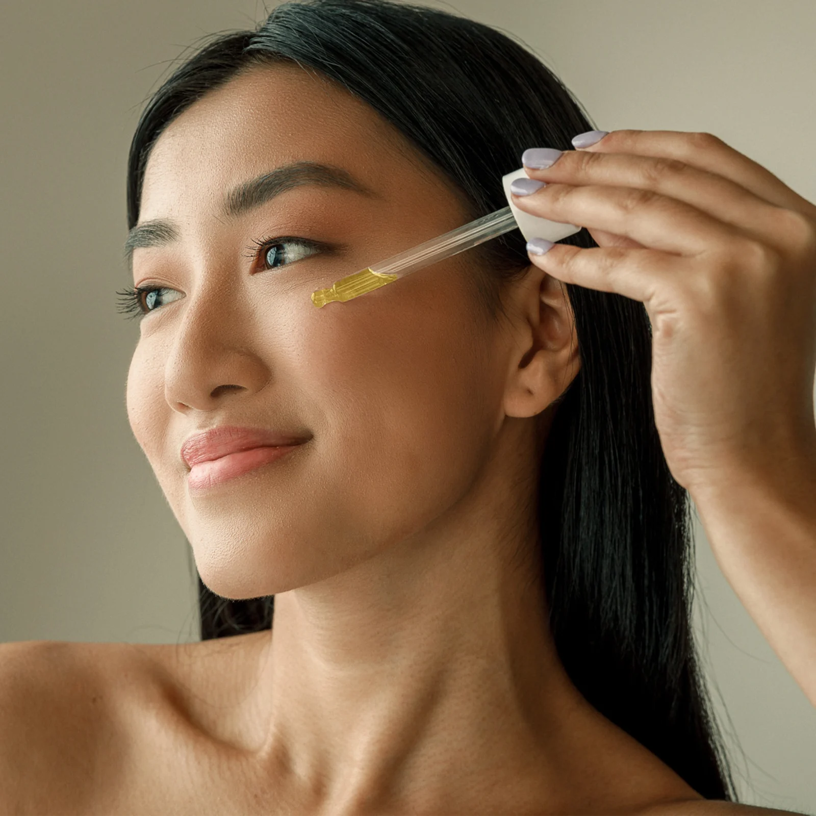 Close-up of a woman receiving a facial treatment with a dropper applying a yellow serum to her temple/cheek area on the side of her face.