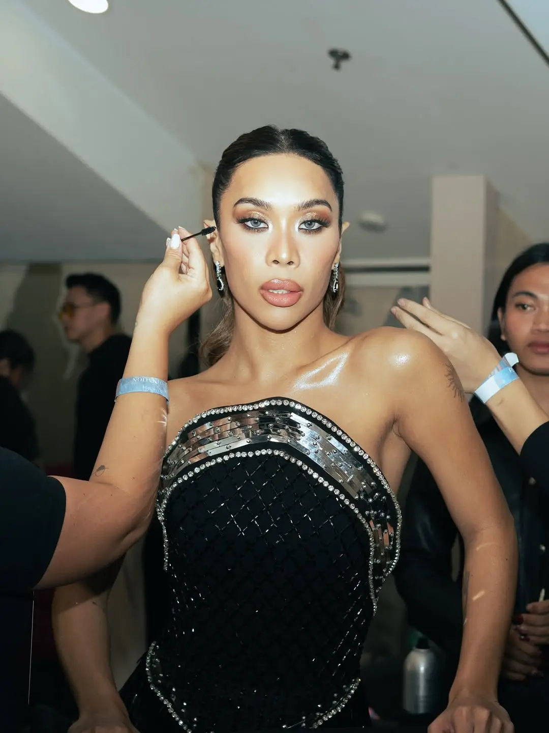 Backstage makeup moment: a woman in a rhinestone-studded black gown has makeup applied by a stylist.