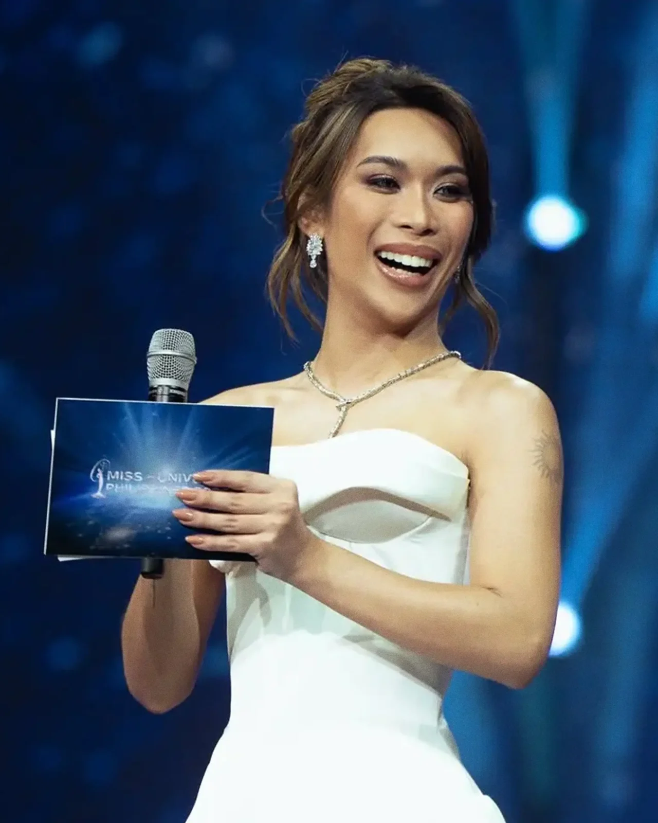 Smiling woman in an off-the-shoulder white dress on stage, holding a blue program and microphone in front of a dark blue backdrop.