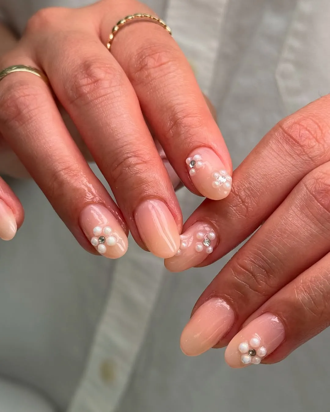 Nude nail design with pearl flower accents on each fingertip, hands with rings visible.