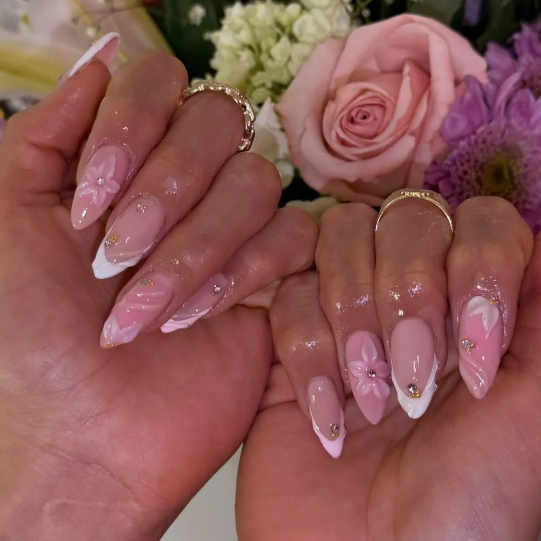 Close-up of hands displaying pink- and white-themed stiletto nail art with 3D pink flowers and rhinestones, glossy finish, bouquet in the background.