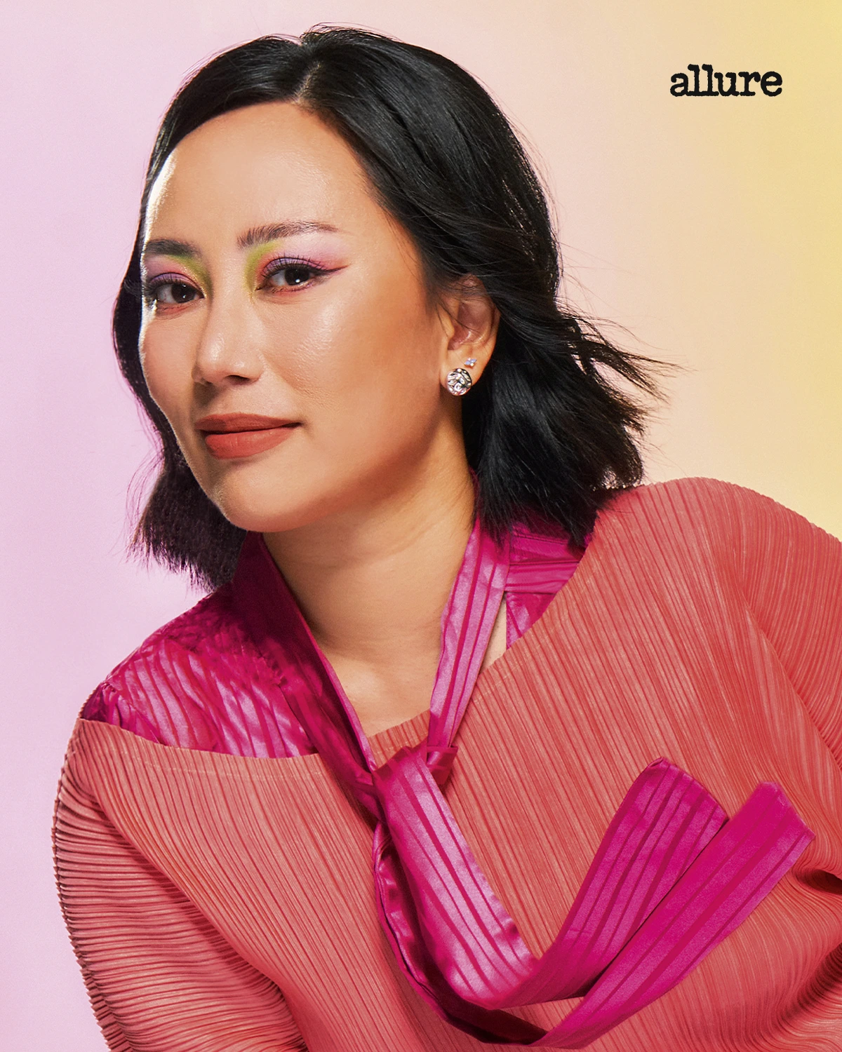 Portrait of a woman with bold pink and green eye makeup wearing a pink pleated outfit against a pastel gradient background; Allure logo in corner.