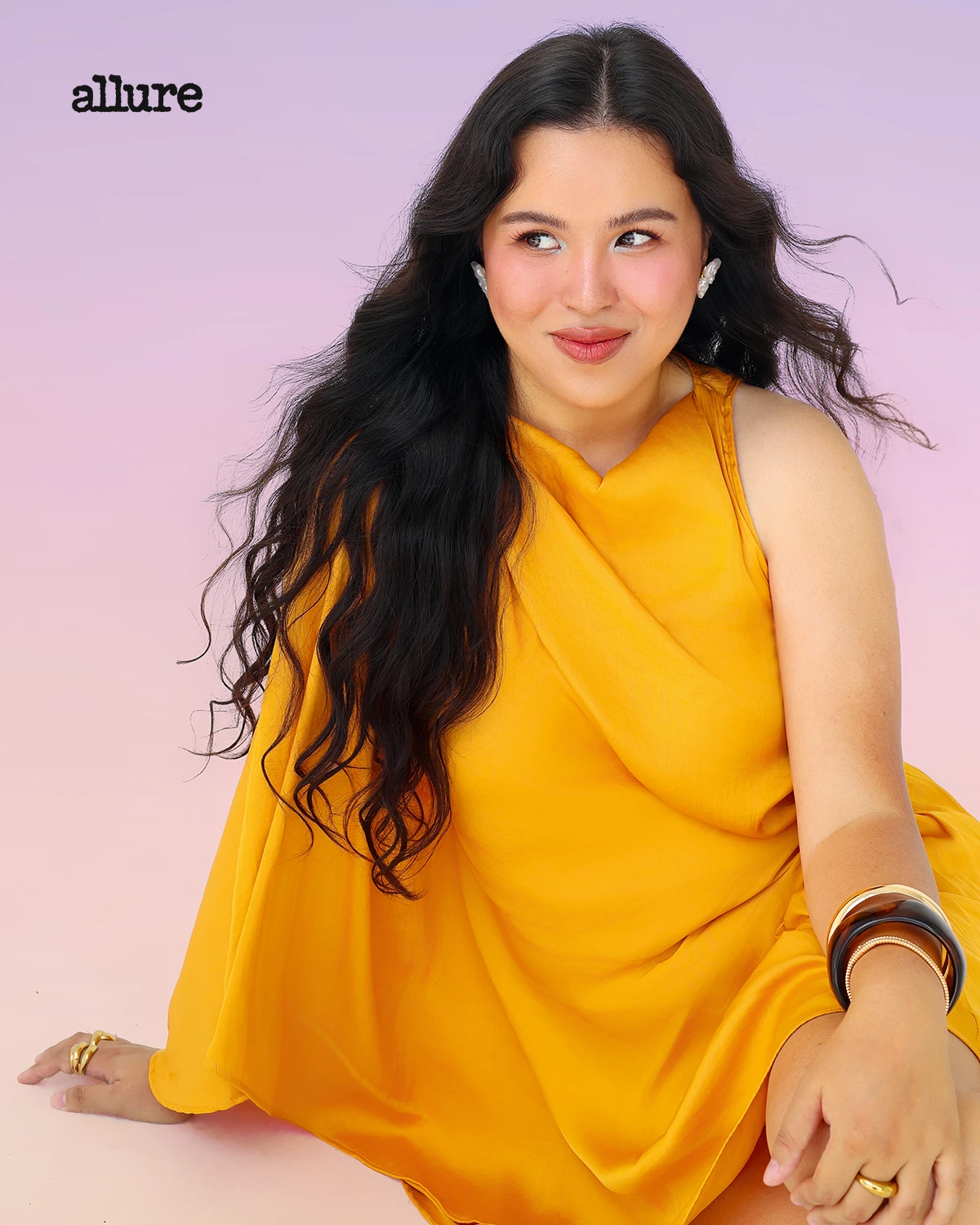 Portrait of a smiling woman with long dark hair in a bright yellow dress, against a pink–lavender gradient background (Allure logo in corner).
