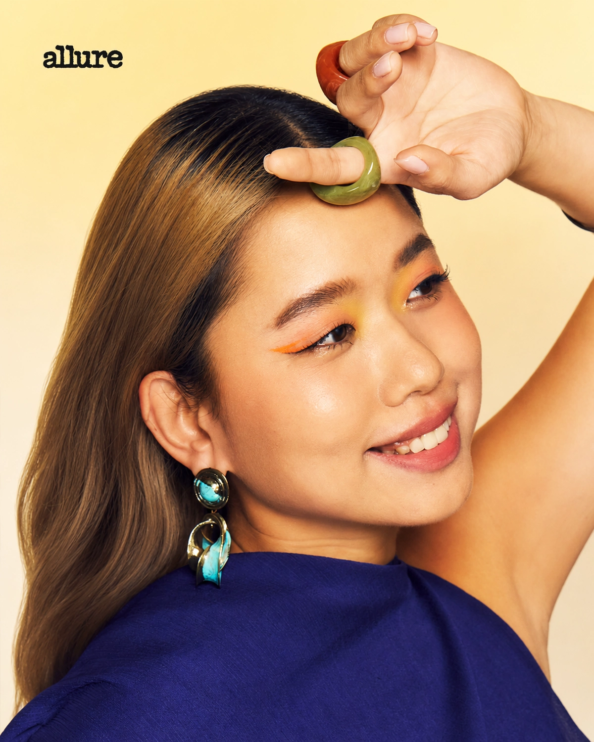 Smiling woman in a blue top with turquoise earrings places a green ring on her forehead; Allure logo in the top-left corner.