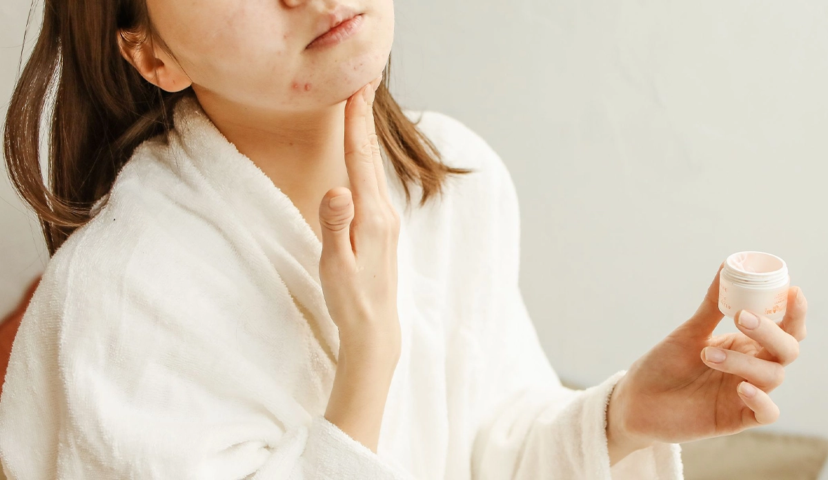 Person in a white bathrobe applying cream to their chin while holding a small cosmetic jar in their other hand, focusing on skincare.