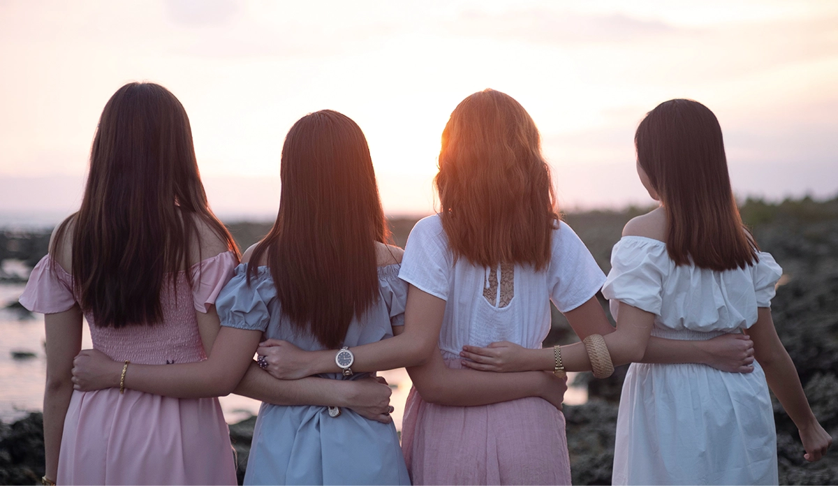 Four women standing arm-in-arm at sunset by a rocky shoreline, seen from behind at the horizon.