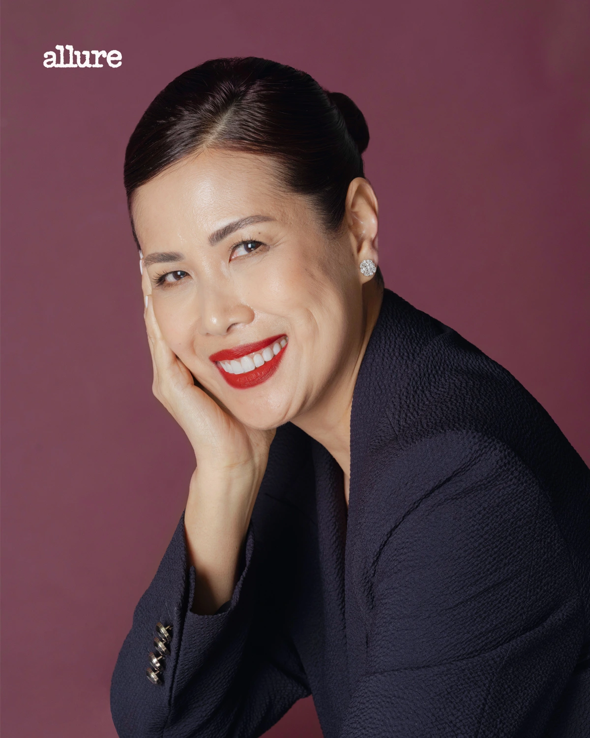 Portrait of a smiling woman with dark hair in a neat bun, red lipstick, and a navy blazer against a burgundy backdrop; Allure logo in the top-left corner.