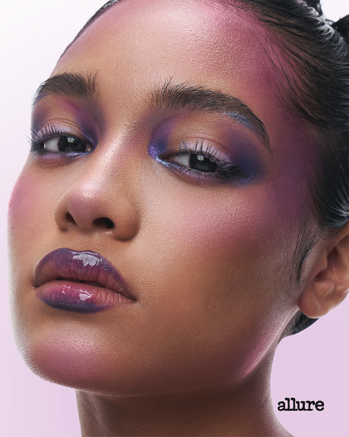 Close-up portrait of a model with bold purple eye makeup, pink blush, and glossy purple lips against a pink background.