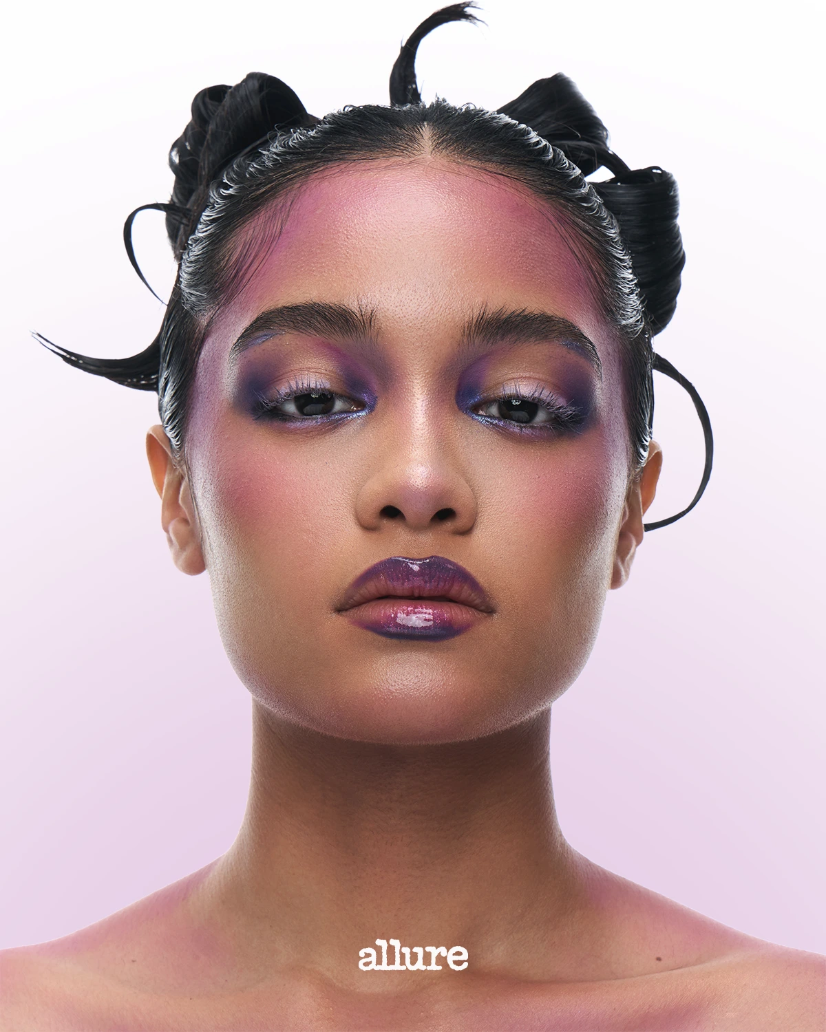 Close-up editorial beauty shot of a model with bold purple-pink eyeshadow, glossy lips, and slicked-back hair against a pale pink background with 'allure' logo at the bottom