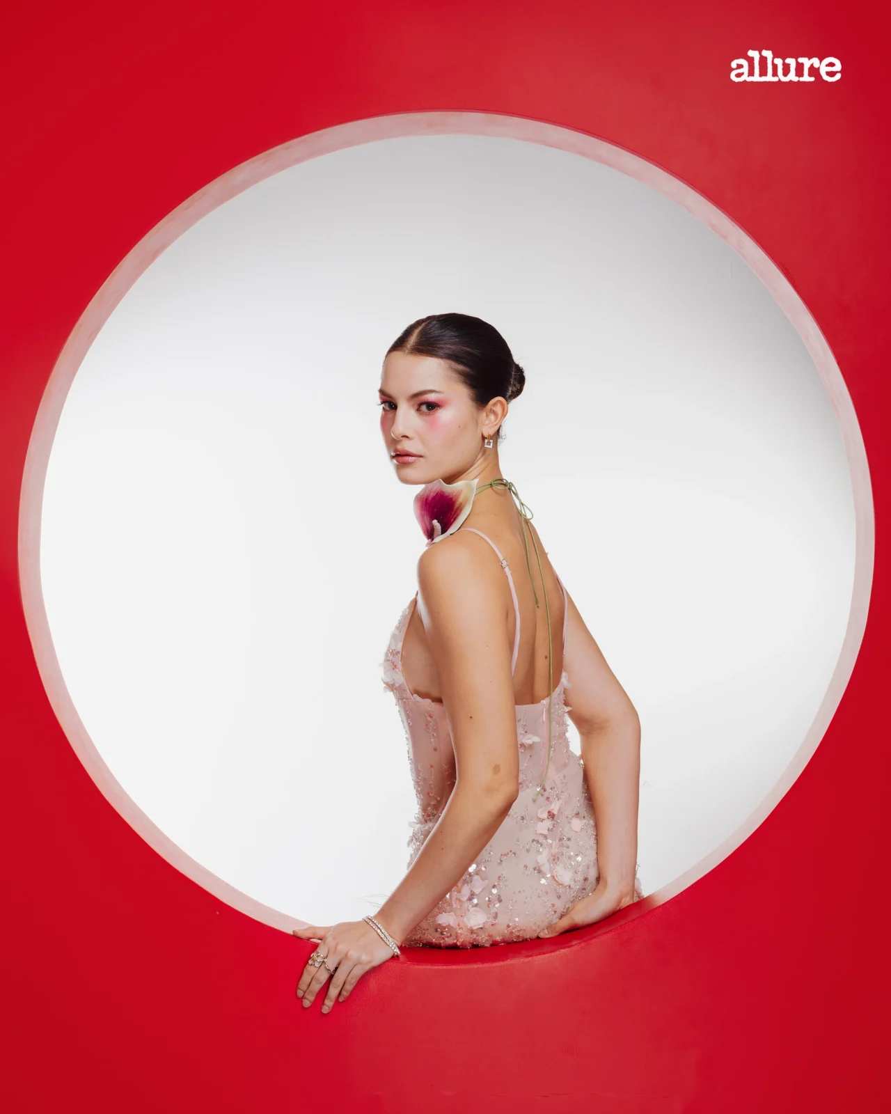 Woman in a pale pink sequin dress sits inside a large white circular frame against a red background, Allure logo in the corner.