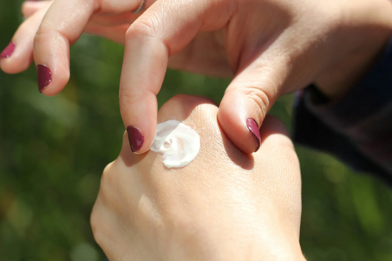 Close-up of hands applying white cream to a child’s cheek outdoors, with maroon-polished nails visible on the fingers