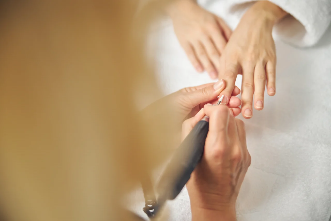 Close-up of hands during a manicure, with a nail technician filing a client's fingernails using an electric nail drill off to the side of the image