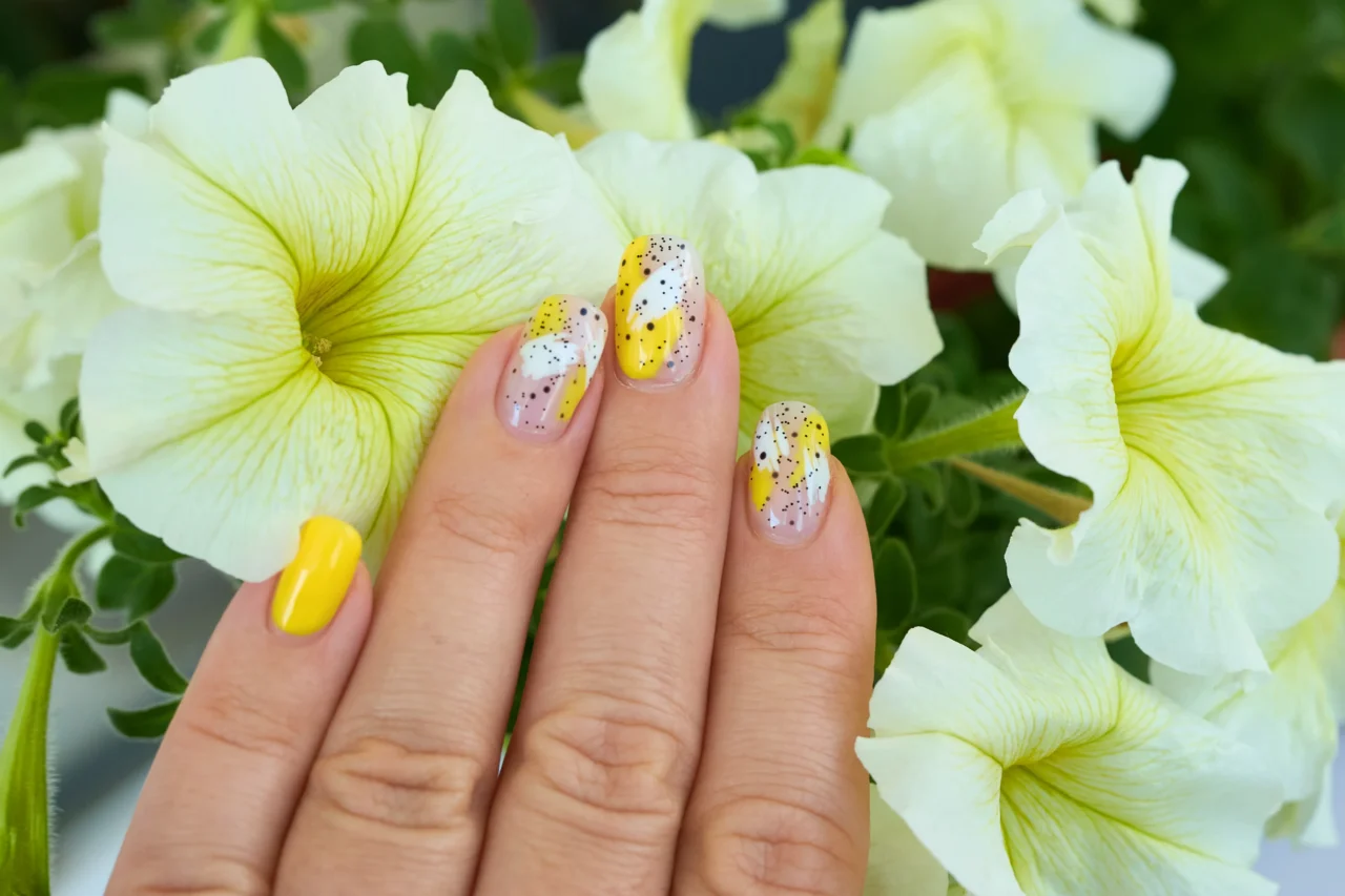 Close-up of manicured nails with yellow and speckled white nail art held against pale yellow hibiscus flowers