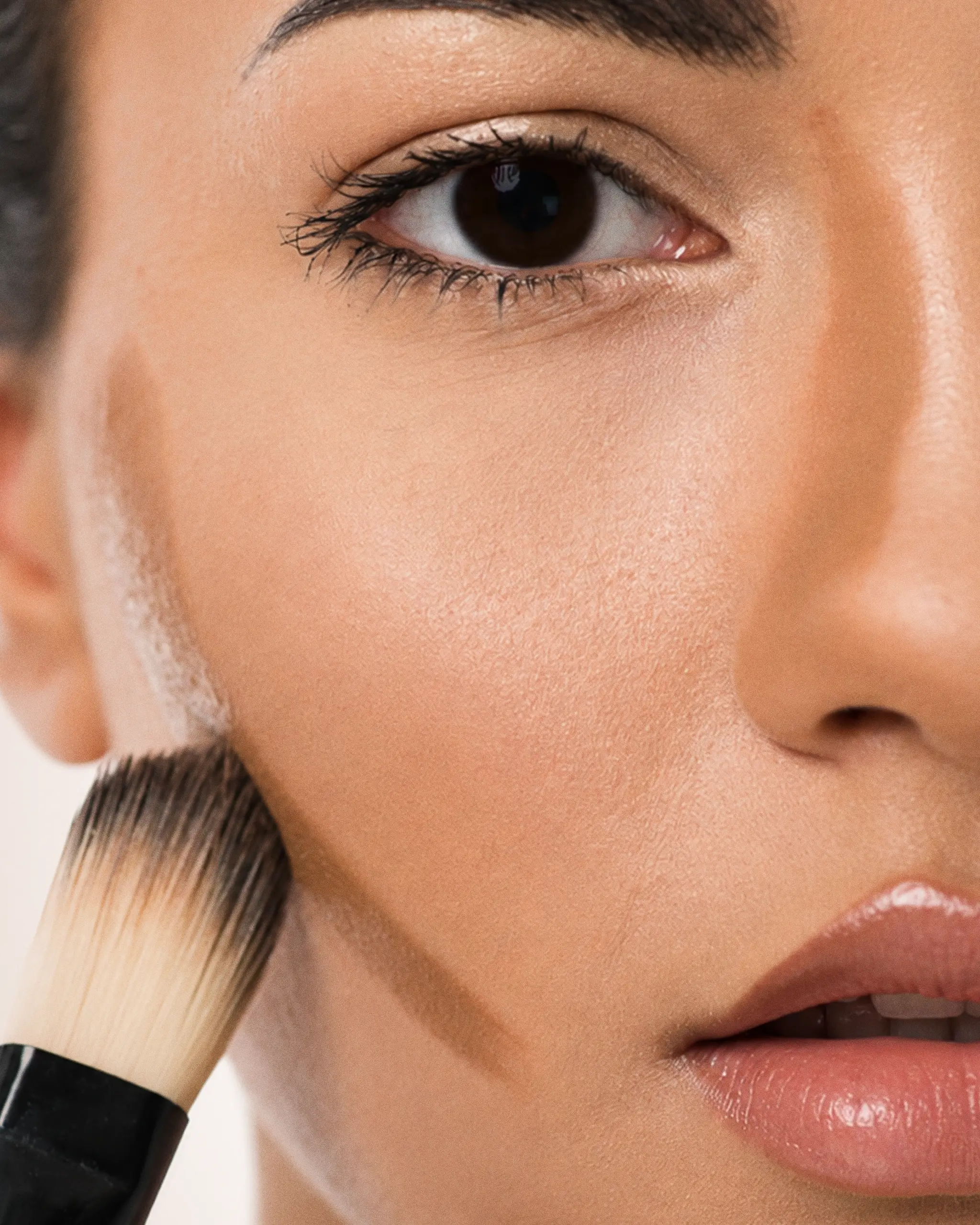 Close-up of a woman's face with a makeup brush touching her cheek, suggesting foundation or powder application.