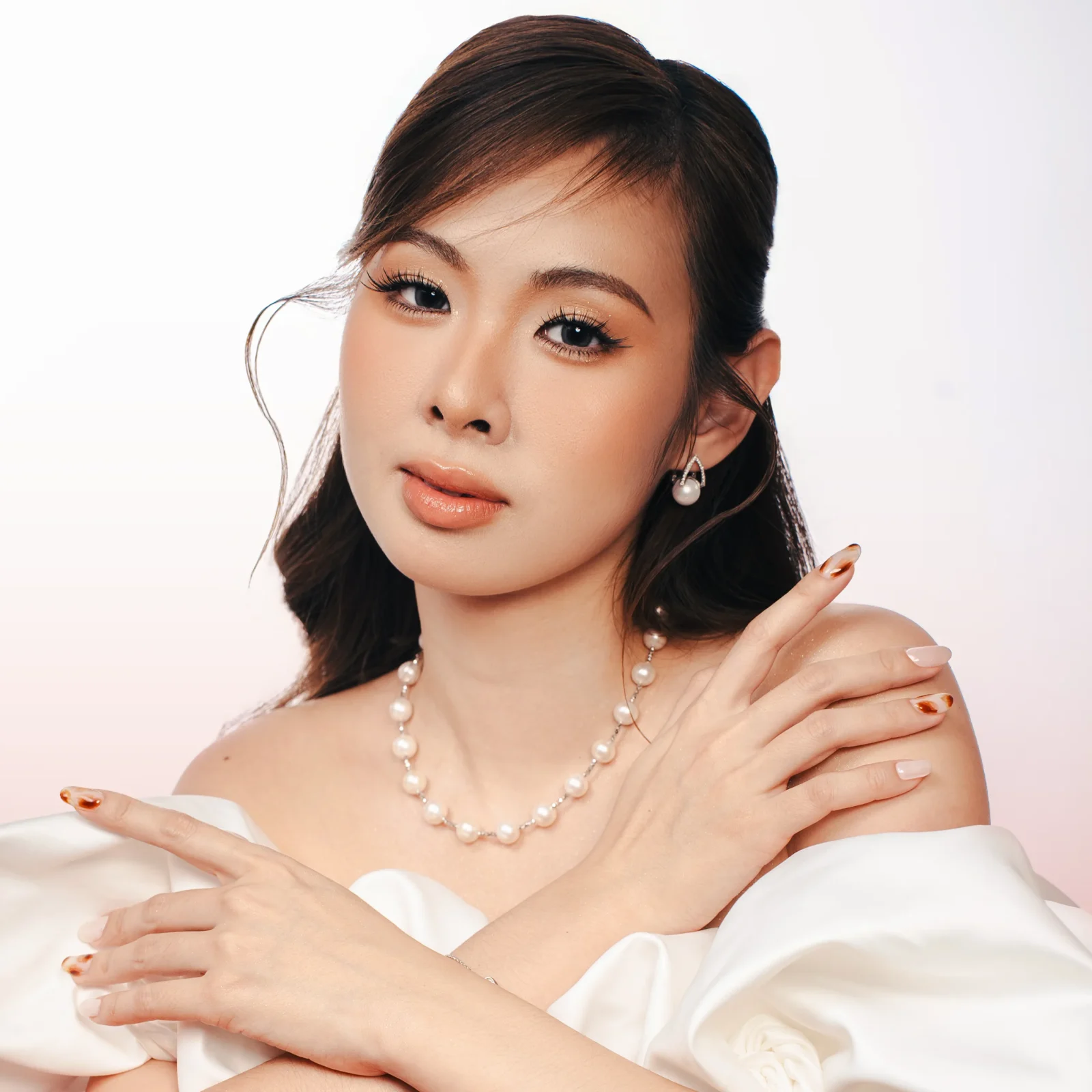 Close-up of a woman wearing pearl jewelry and an off-shoulder white dress, with soft makeup and a calm expression.