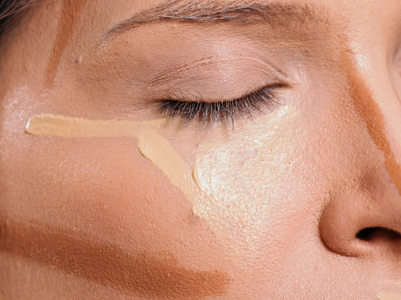 Close-up of a closed eyelid with beige makeup and mascara on the lashes, showing skin texture.