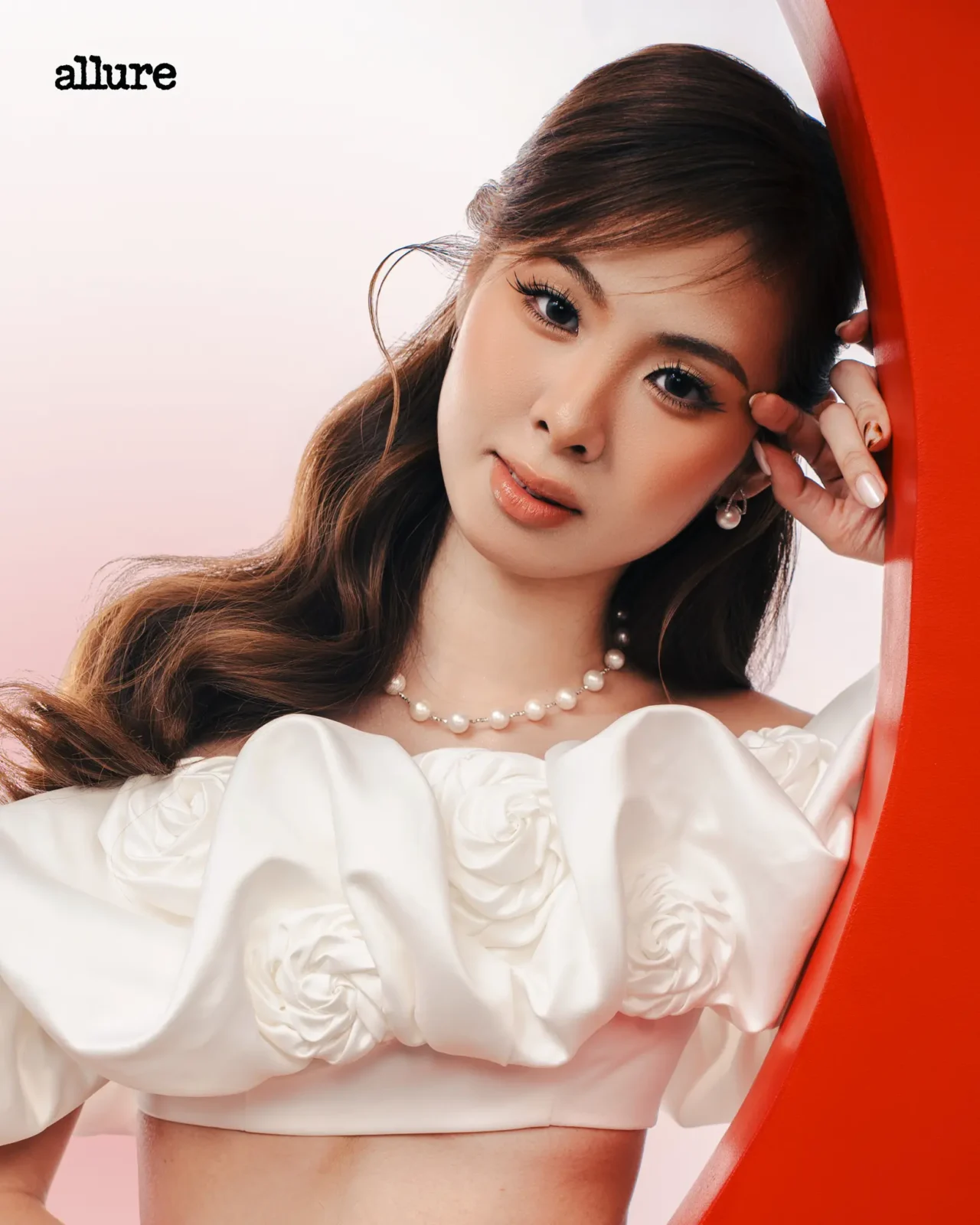 Portrait of a woman in a white ruffled top with a pearl necklace, leaning against an orange curved surface; Allure logo visible.