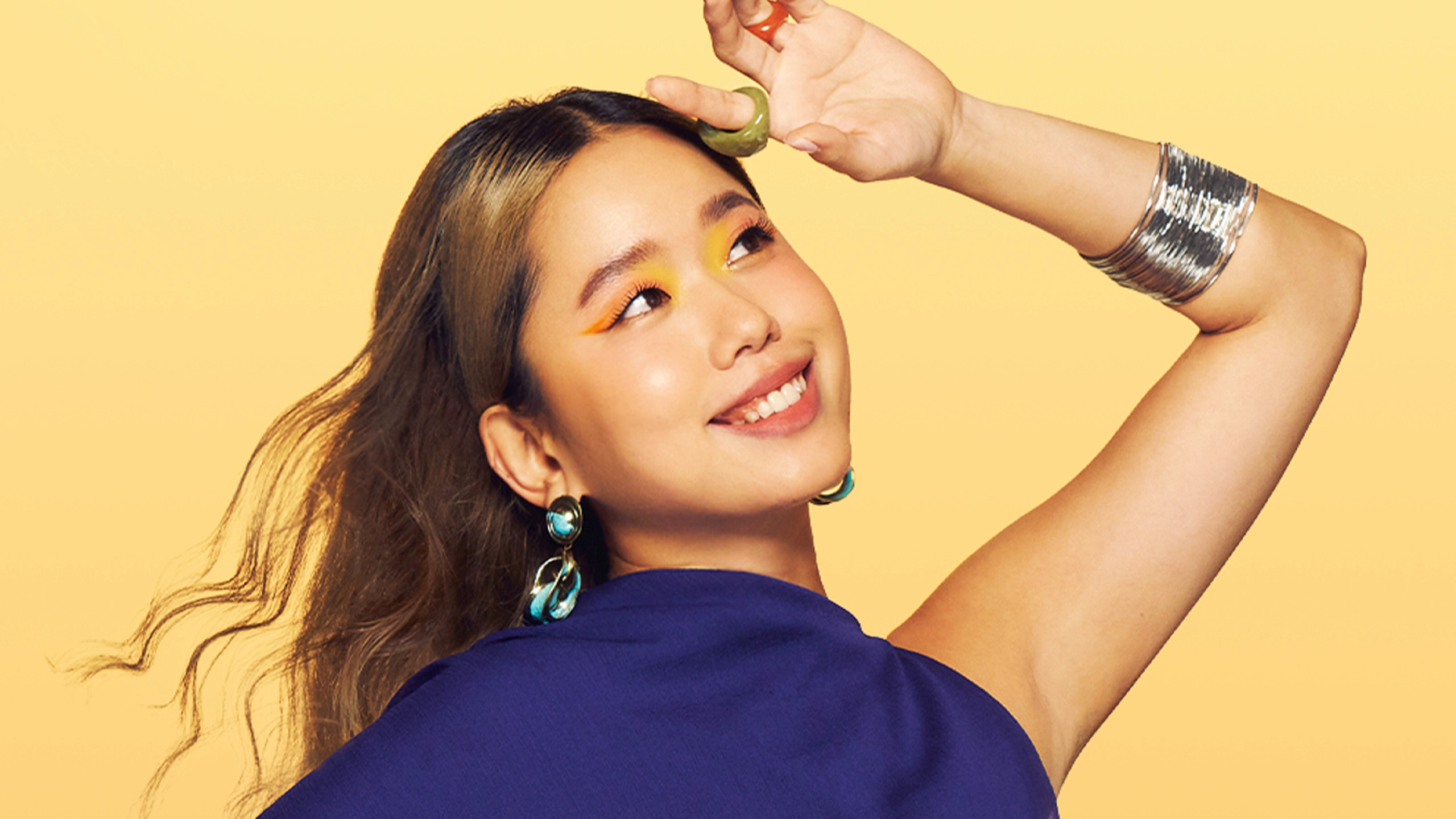 Smiling woman in a blue top wearing turquoise earrings and silver bracelets, balancing a green ring on her hair against a yellow backdrop.