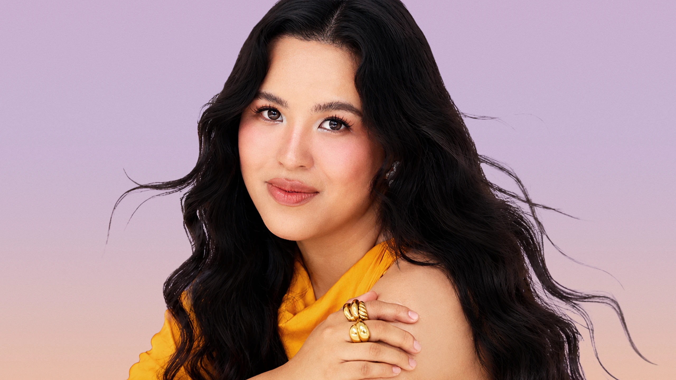Portrait of a woman with long dark hair wearing an orange top, looking at the camera against a soft pink–lavender gradient background.