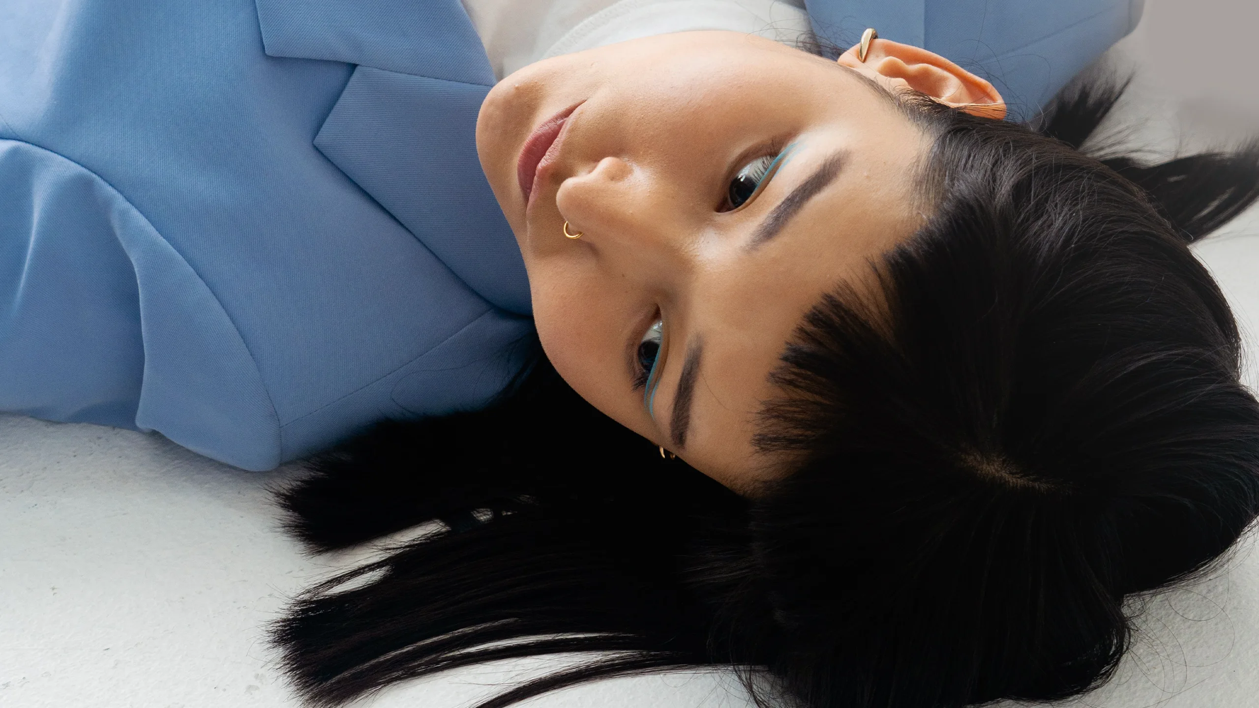 Person lying on a white surface with head turned to the side, wearing a blue outfit and turquoise eye makeup with a gold hoop earring