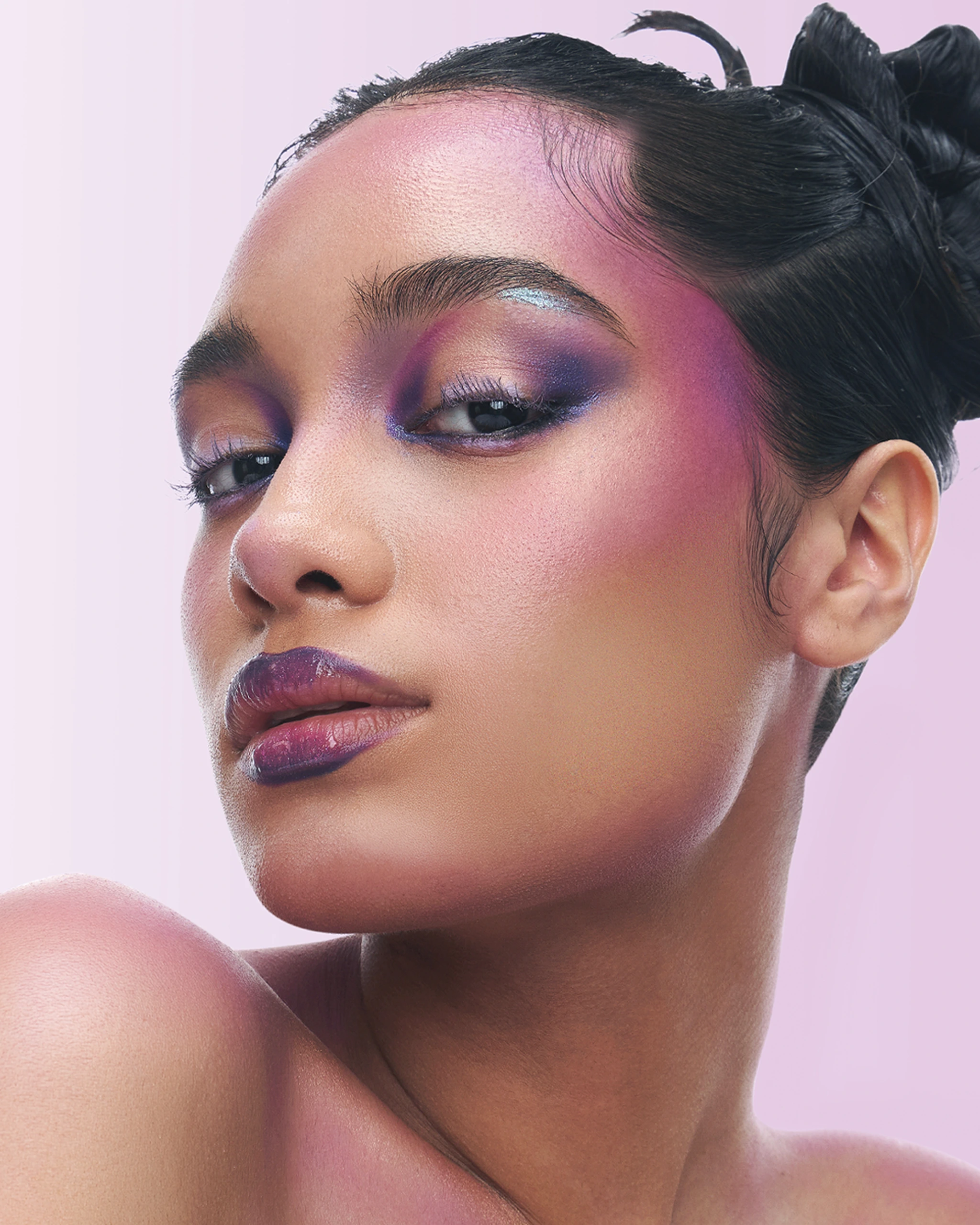 Close-up of a woman's face with bold pink eyeshadow and purple eye makeup against a pale pink background, lips tinted pink.