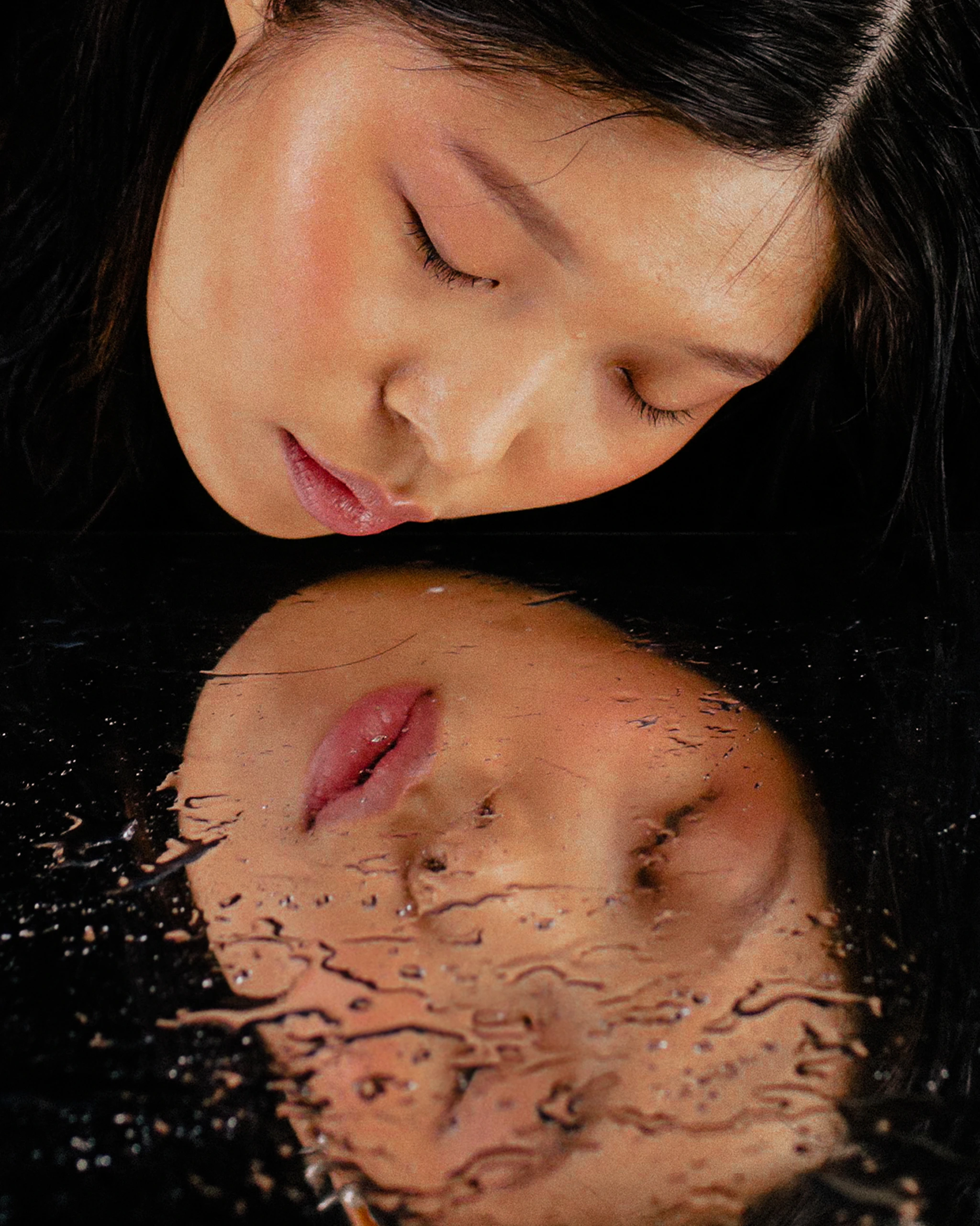 Close-up of a woman resting her face on a dark reflective surface, eyes closed, creating a mirror image below.